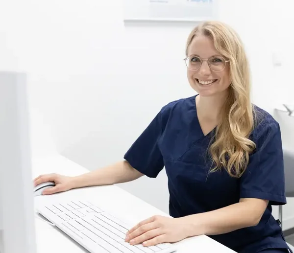 Eine Frau in blauer Arbeitskleidung sitzt an einem Schreibtisch vor einem Computer, hält eine Hand auf der Maus und lächelt in die Kamera.