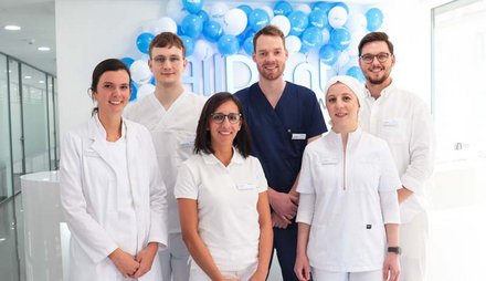 Sechs Personen in medizinischer Arbeitskleidung stehen vor einer Wand mit blauen und weißen Luftballons.