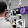 Zahnarzt arbeitet mit einem Dentalmikroskop, w&auml;hrend auf Monitoren vergr&ouml;&szlig;erte Aufnahmen eines Zahns und R&ouml;ntgenbilder zu sehen sind