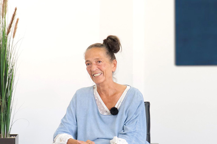 Frau mit Dutt und blauem Pullover sitzt vor weißer Wand, links steht eine Pflanze